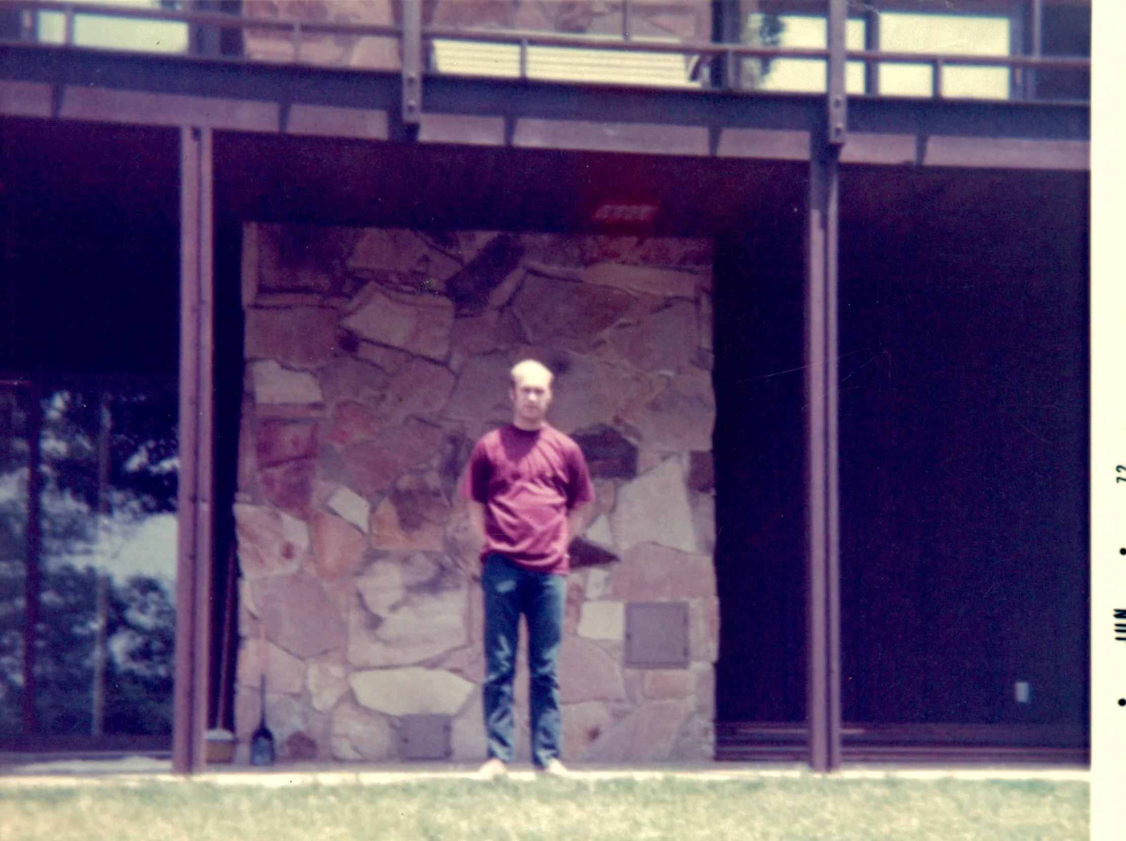 <strong>Charlie Eesley standing in front of the stone fireplace at Highland Ridge.</strong> Confirmed by Chuck Eesley 2026. <strong>Charles 'Charlie' McMaster Eesley</strong> — Chuck's father — casually posed in red shirt and jeans in front of the stone fireplace of the house his architect father Will designed. The deck above is visible, the stone hearth at center, the large multi-paned windows on the right. <strong>Charlie at home in his father's architecture — the house he inherited and raised his own son in.</strong> The earlier hypothesis that this was Will himself is withdrawn.