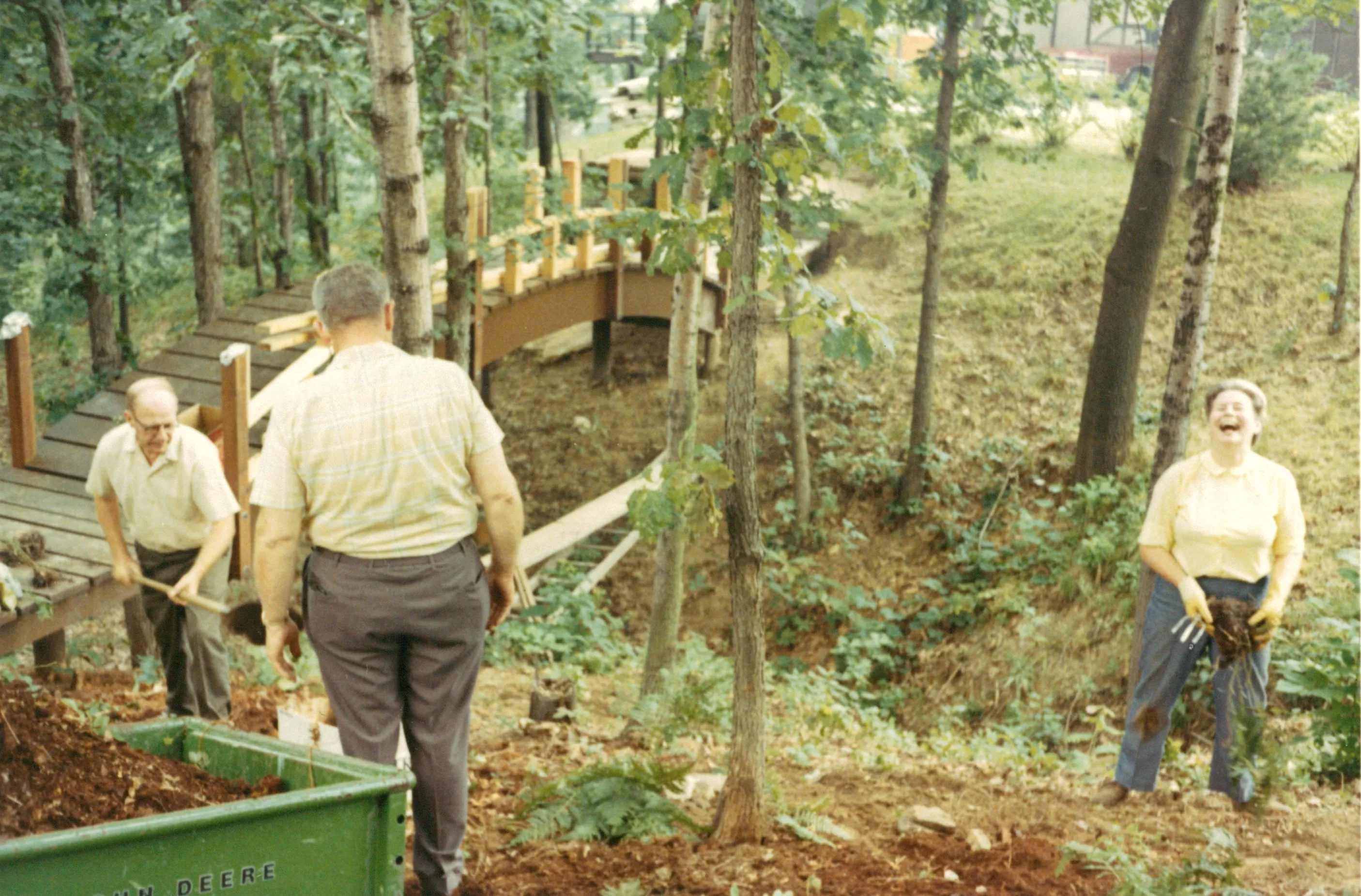 <strong>The red bridge being worked on. Will Eesley with shovel.</strong> Confirmed by Chuck Eesley 2026. <strong>Will Eesley</strong> at left in red shirt with shovel — building or repairing the bridge he designed. The two other figures (man with back to camera in middle in plaid trousers, laughing woman in yellow at right beside the green 'JOHN DEERE' wheelbarrow) are open for identification. The bridge in the background has its railings up but appears in mid-construction or mid-repair. The labor side of the property — the orchard, the bridge, the firewood — the eulogy named explicitly.