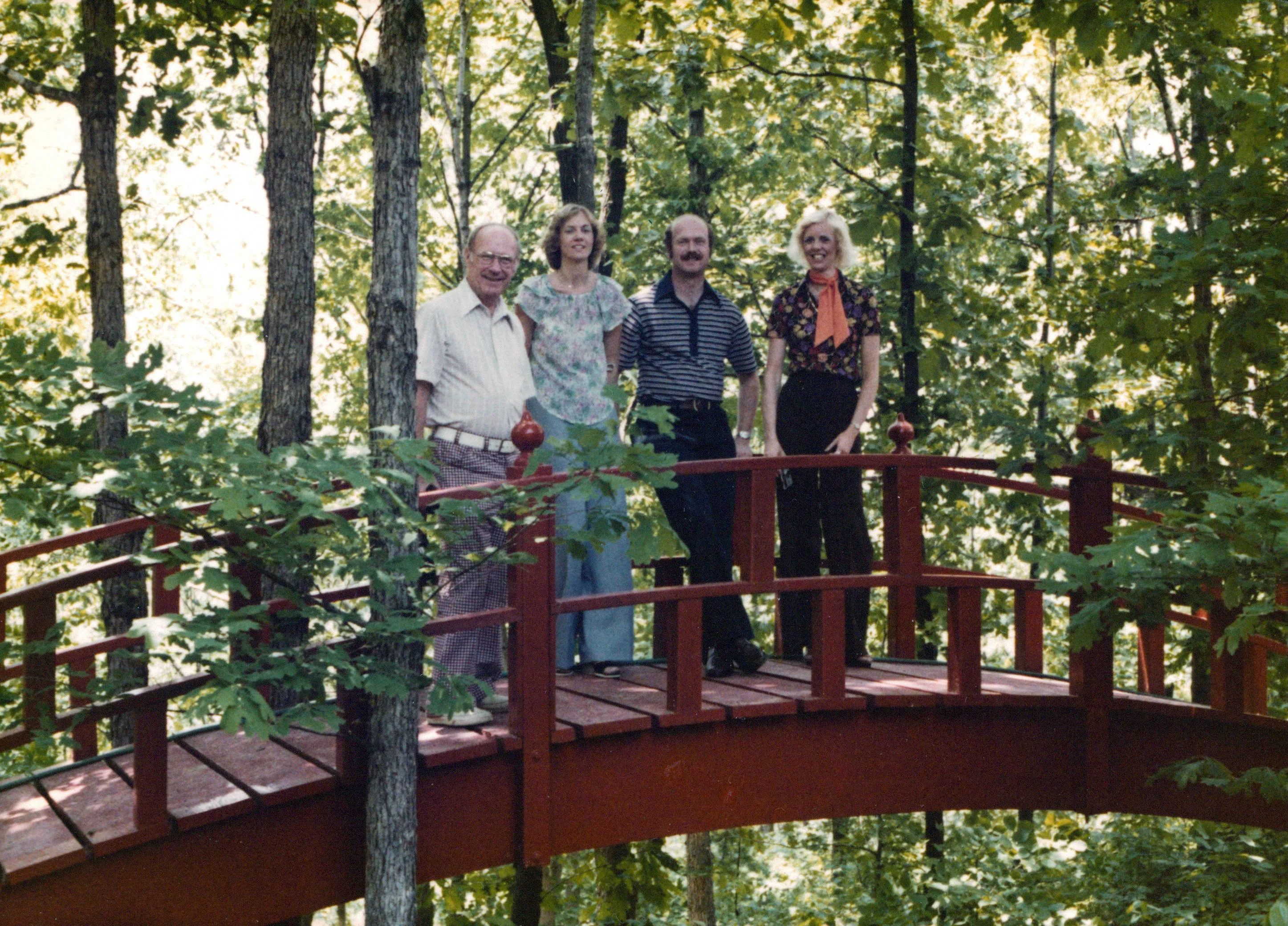 <strong>Four people on the small red bridge — Will Eesley, Charlie, Terrie, and Aunt Sandy.</strong> Confirmed by Chuck Eesley 2026. <strong>Will Eesley</strong> the architect at left in plaid trousers and white shirt, on the bridge he himself built. <strong>Terrie Lee Wildermuth Eesley</strong> (Chuck's mother) beside him in pale floral blouse and dark pants. <strong>Charles 'Charlie' Eesley</strong> with mustache in dark patterned shirt — Will's son, Terrie's husband, Chuck's father. <strong>Sandra Sue (Wildermuth) Clement</strong> — 'Aunt Sandy,' Terrie's younger sister and Robert Earl's second daughter — at far right in dark vest and orange neckscarf. The photograph captures a small but consequential geography: <strong>the bridge on the paternal Eesley grandfather's property hosting both sides of Chuck's parents' families at once.</strong>