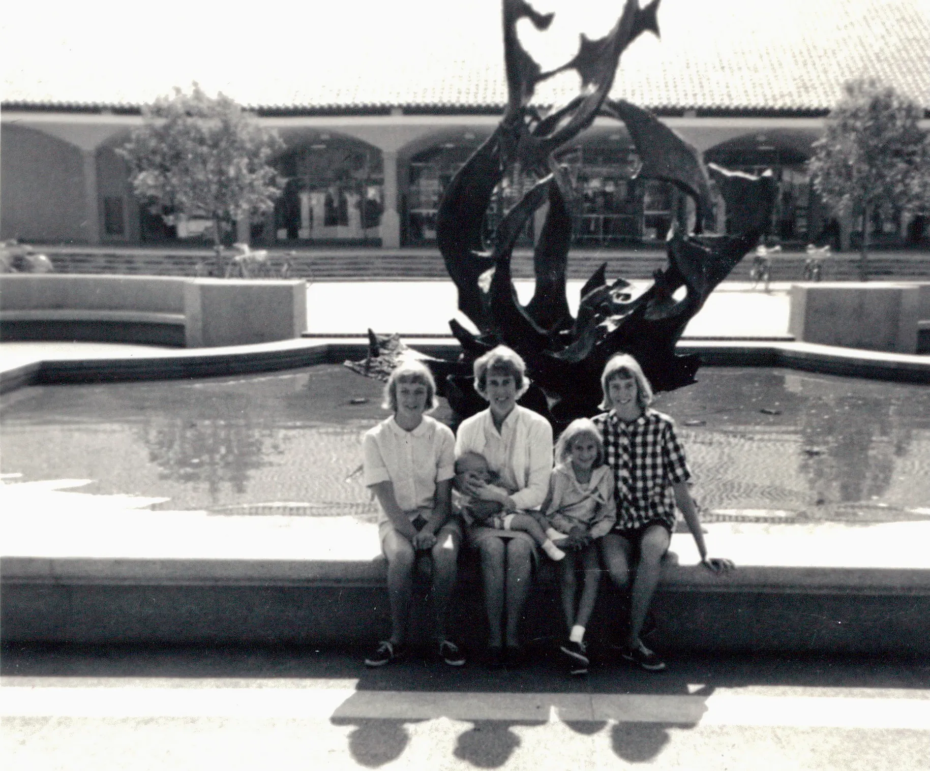 Family at the White Memorial Fountain (the bronze 'Claw' by Aristides Demetrios, installed only a year earlier in 1964) in the heart of campus, on **24 August 1965**. Identified by Chuck: Uncle Rob Wildermuth, Chuck's mother Terrie Lee (then about sixteen), Sandy (Sandra Sue Wildermuth Clement), and Debbie Wildermuth, with their mother Dorothy 'Dottie' Davis Wildermuth holding the youngest. Both the **original black-and-white print** and Chuck's **AI-colorized rendering** are below — the B&W is the documentary record; the colorization is an interpretive aid for picturing the day.
