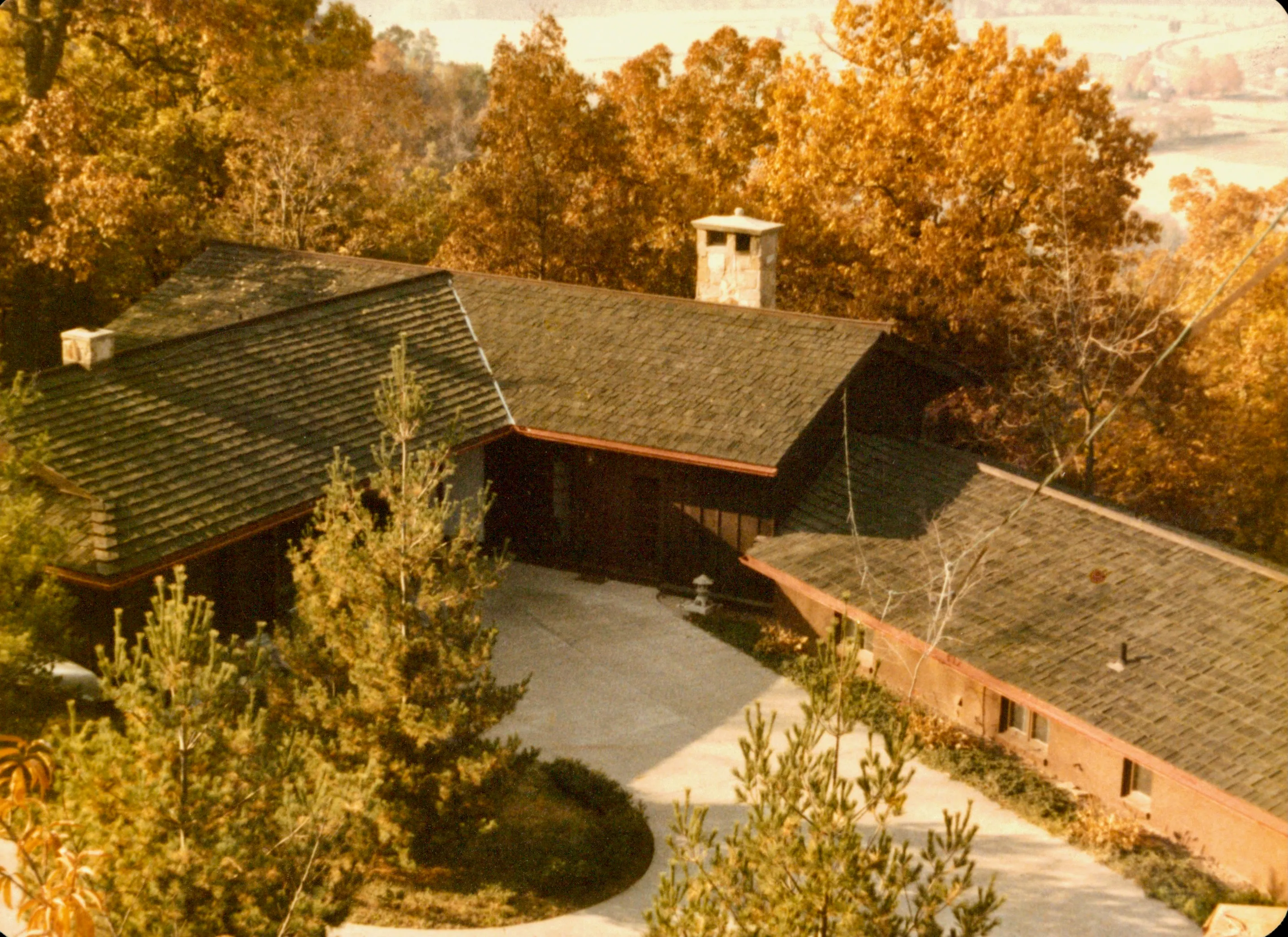 <strong>The house from above, fall colors.</strong> Will's design visible in plan — the L-shaped wood-shingle roof, the large central chimney, the driveway circling the house, and the orchard trees Will planted dotting the property. Taken from elevation behind the house, possibly the upper ridge. The colors fit late October.