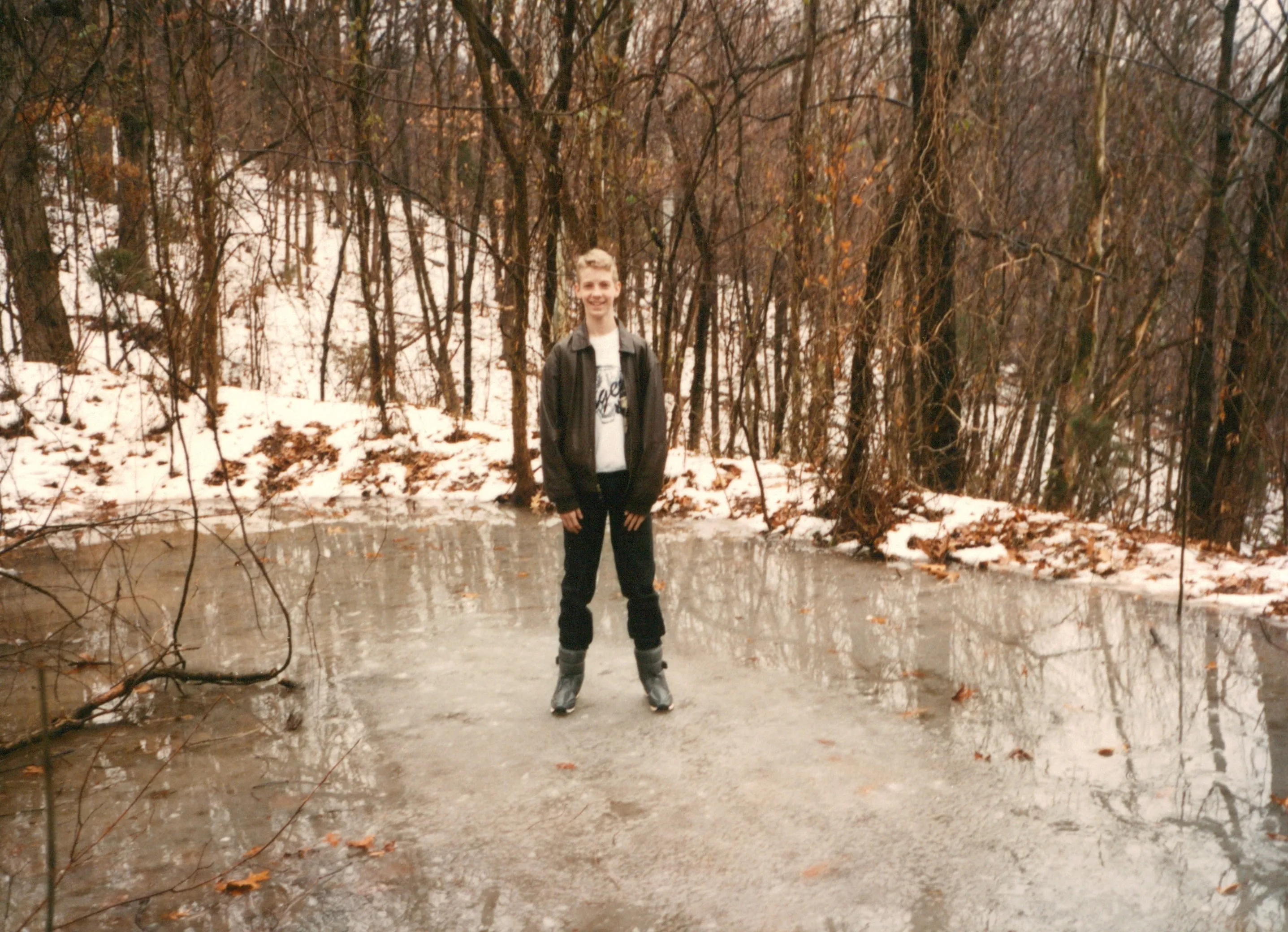 <strong>Young Chuck on the frozen pond at Highland Ridge.</strong> Confirmed by Chuck Eesley 2026. <strong>Young Chuck Eesley</strong> — about 12–15 — standing on the ice in dark pants and brown jacket, in winter, woods bare behind. <strong>This is the pond his father's <a href='/docs/eulogy-charles-mcmaster-eesley/'>eulogy</a> describes him breaking through as a smaller boy</strong> — the pond Charlie pulled him out of, his boots staying behind 'still stuck in the mud on the bottom to this day.' The photograph captures Chuck a few years older, the ice frozen solidly enough now to bear him, the boots presumably still in the mud below the surface he's standing on.