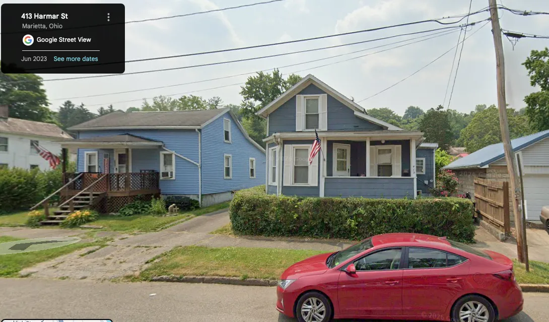 Google Street View of 413 Harmar Street, Marietta, c. 2026 — the Wildermuth family house still standing eighty-plus years after Earl Adam and Sadye Irene bought it in 1929. Blue clapboard two-story house with a covered front porch, brick foundation visible at the base (the same brick foundation Robert Earl's 1989 memoir describes as 'approximately four feet high'), an American flag hanging from a porch column, and the street's 'steady rise' visible in the background hills of the West Side of Marietta. The house, the West-Side neighborhood, and the topography of Robert Earl's childhood are all still on the ground a century after Earl Adam bought the house at the bottom of the Depression in 1929.