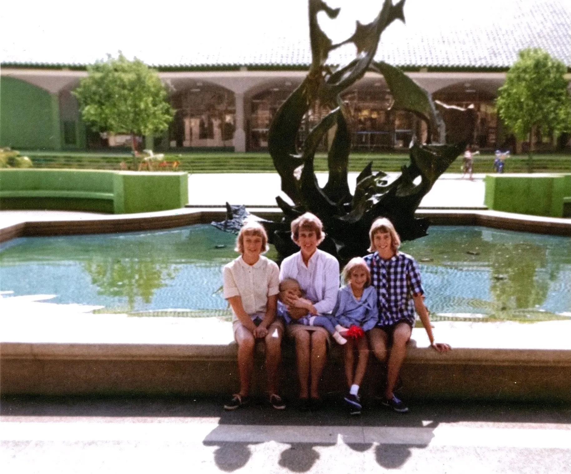 Family at the White Memorial Fountain (the bronze 'Claw' by Aristides Demetrios, installed only a year earlier in 1964) in the heart of campus, on **24 August 1965**. Identified by Chuck: Uncle Rob Wildermuth, Chuck's mother Terrie Lee (then about sixteen), Sandy (Sandra Sue Wildermuth Clement), and Debbie Wildermuth, with their mother Dorothy 'Dottie' Davis Wildermuth holding the youngest. Both the **original black-and-white print** and Chuck's **AI-colorized rendering** are below — the B&W is the documentary record; the colorization is an interpretive aid for picturing the day.