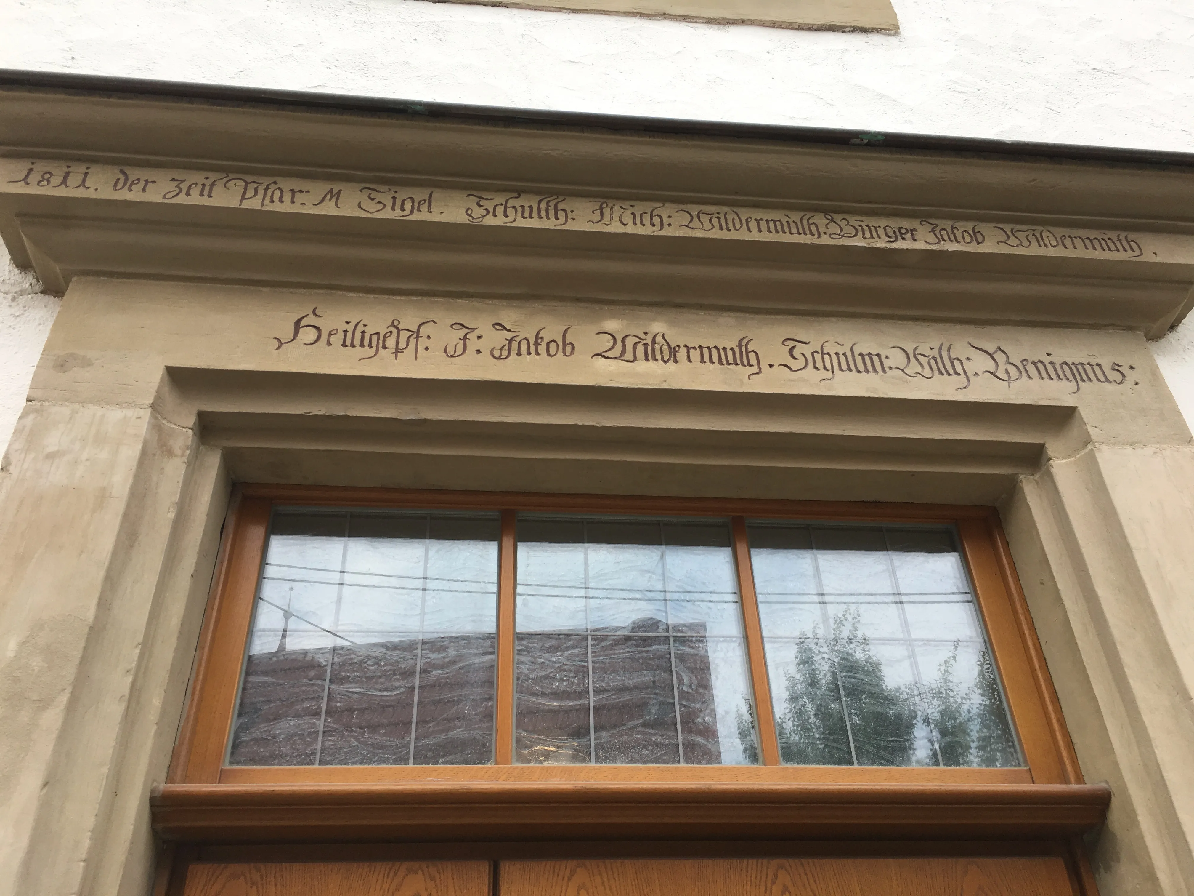 The 1811 stone door-lintel inscription on one of the square's civic buildings: three Wildermuths named as Schultheiß (Michael), Bürger (Jakob), and Heiligenpfleger (J. Jakob), alongside the schoolmaster Wilhelm Benignus and the pastor M. Sigel.