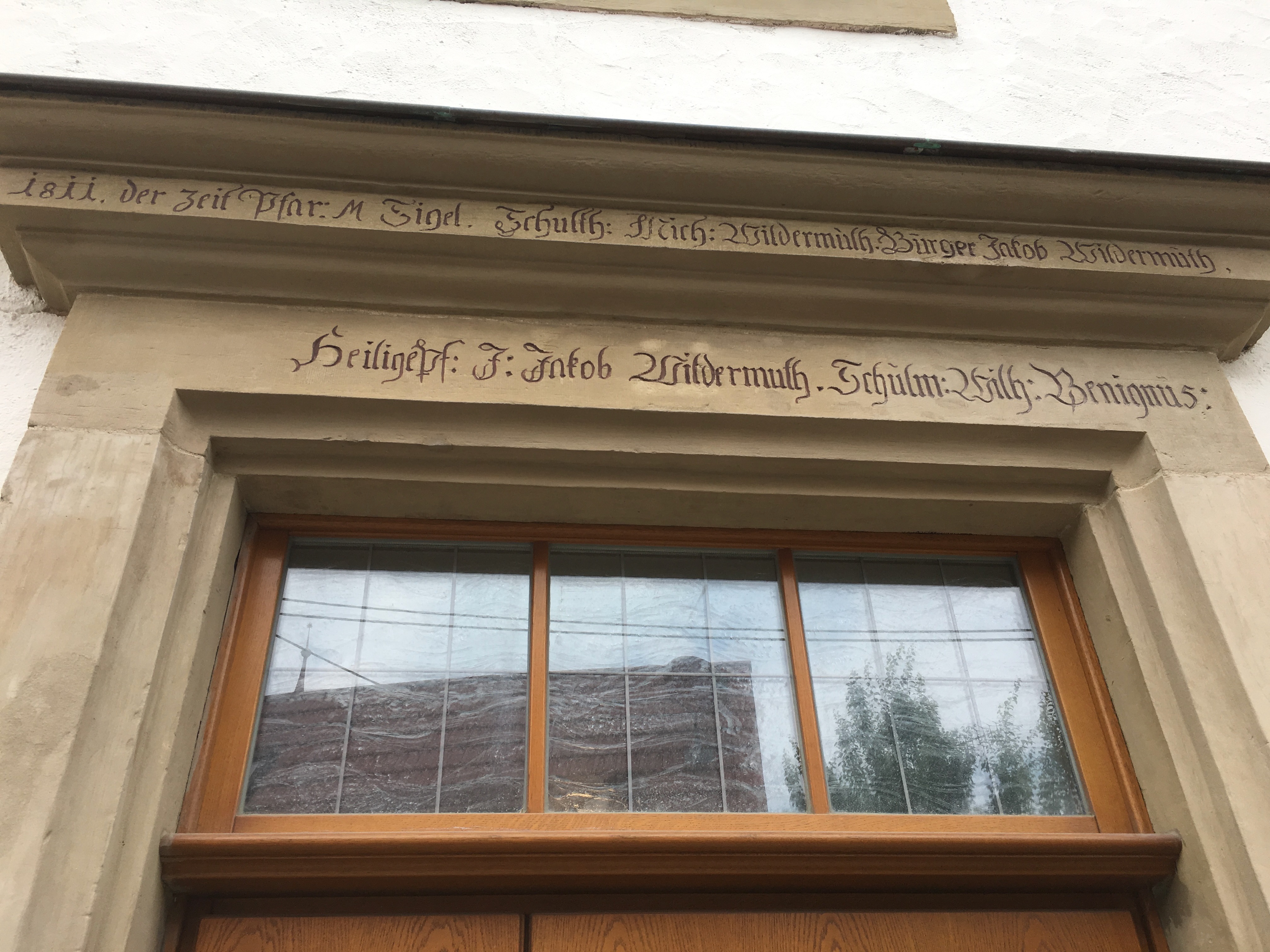 The 1811 stone door-lintel inscription on one of the square's civic buildings: three Wildermuths named as Schultheiß (Michael), Bürger (Jakob), and Heiligenpfleger (J. Jakob), alongside the schoolmaster Wilhelm Benignus and the pastor M. Sigel.