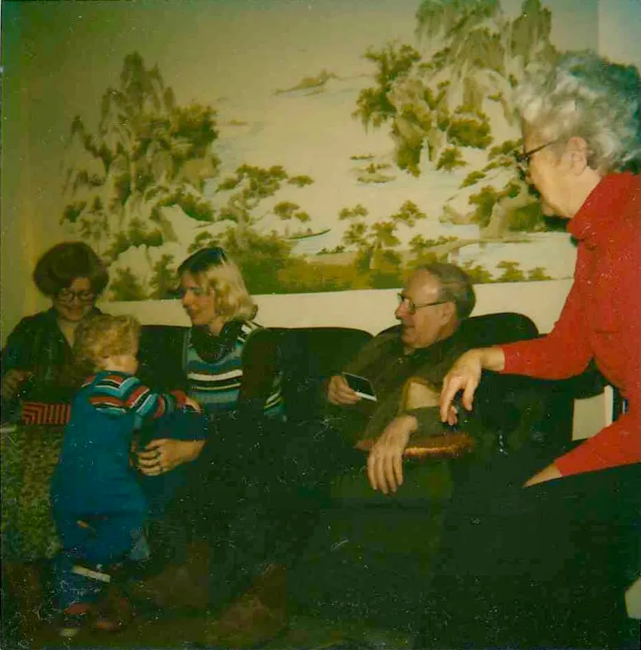 <strong>Young Chuck and the Chinese mural at Highland Ridge — with Grandpa Will Eesley, Grandma Peggy Eesley, Aunt Jeanne, and his mother Terrie.</strong> Full cast confirmed by Chuck Eesley June 2026. Young Chuck — about one to two and a half years old — walking on the floor in colorful striped overalls in front of the family couch, with the large Chinese mountain-and-river landscape mural behind it that decorated one wall of the Highland Ridge living room. Watching him: <strong>Aunt Jeanne (Eesley) Kamiab</strong> on the left; <strong>Will Eesley</strong> the architect-grandfather on the right with glasses, in the house he himself designed; <strong>Grandma Peggy McMaster Eesley</strong> beside him in red; and <strong>Terrie Lee (Wildermuth) Eesley</strong> — Chuck's mother — seated, blonde, in the striped sweater. Both paternal Eesley grandparents in the frame, in the architect-grandfather's own East-Asian-inspired living room, with the Chinese mural behind him — almost certainly a souvenir of the Japan / Hong Kong travels that informed his architecture. The Chinese mountain-and-river landscape mural is a distinctive interior feature of the Highland Ridge house, locating the photograph specifically there.