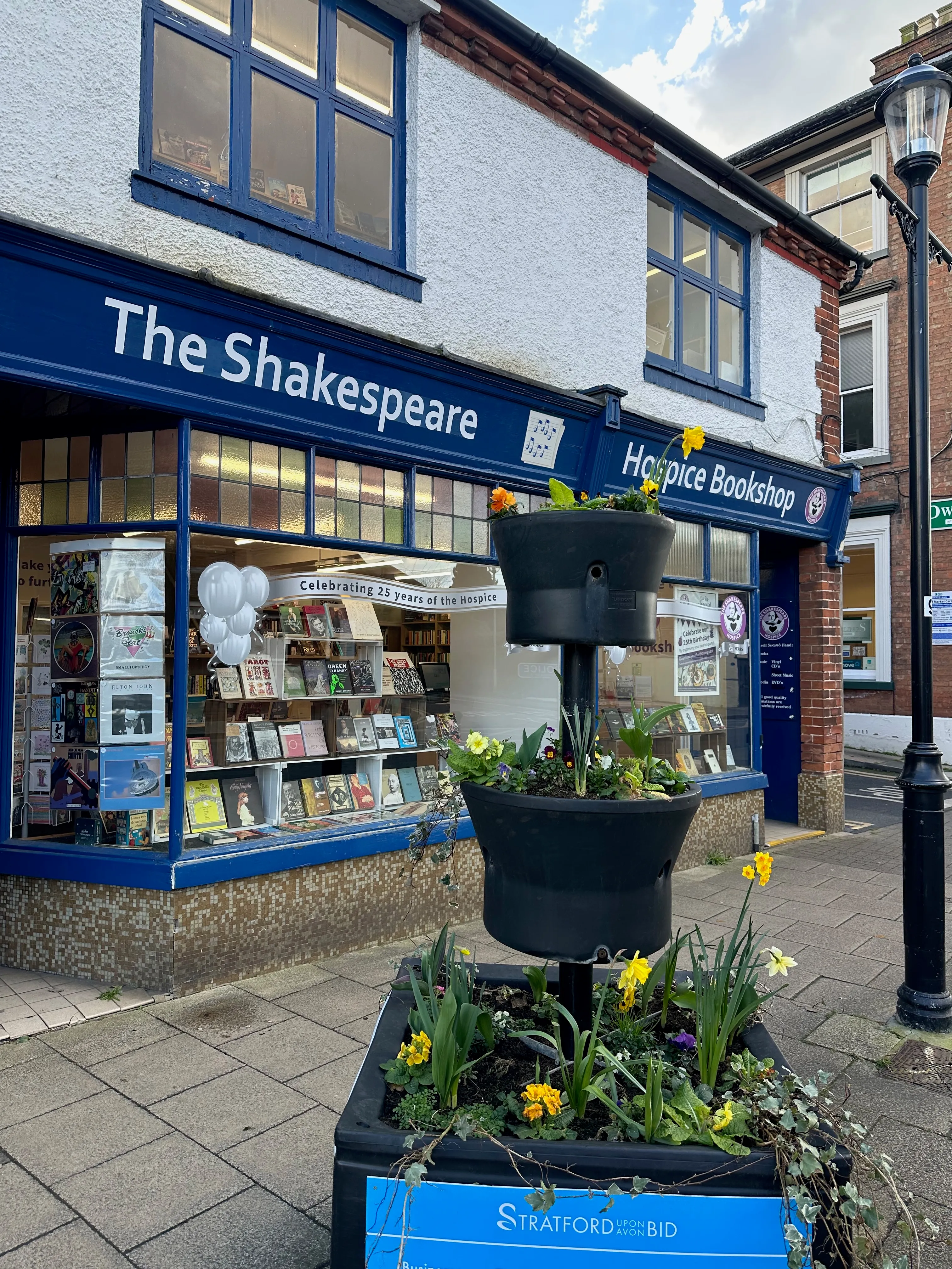 Chuck and Lijie at the building during their England research trip. The shopfront now reads 'The Shakespeare Hospice Bookshop' — a charity bookshop, hospice-affiliated, in the old building.
