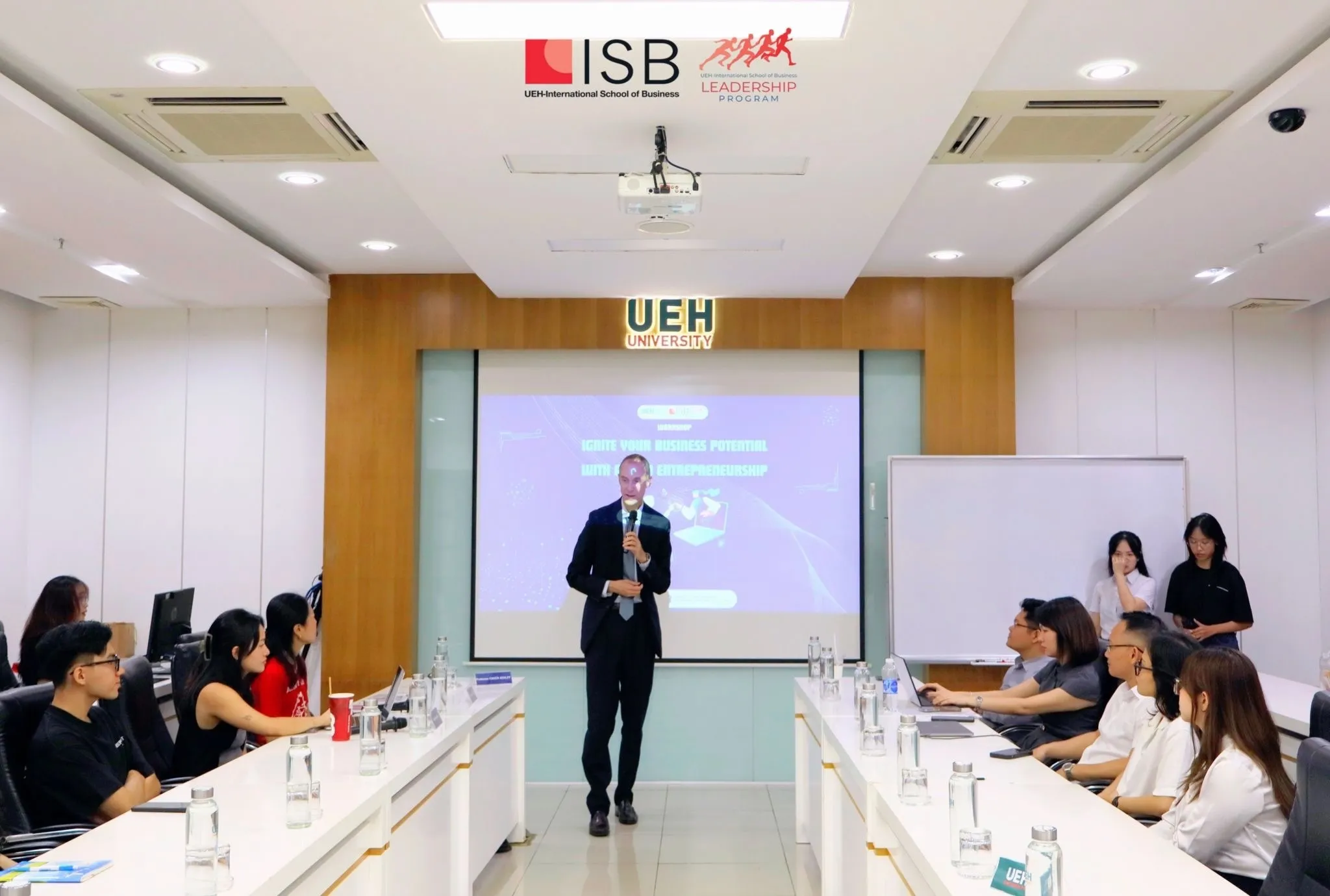 Chuck has returned to Ho Chi Minh City multiple times in the years since the June 2011 first visit, including teaching trips at local universities with the family foundation. The photograph shows Chuck giving a December 2024 presentation in a lecture hall at the **UEH-International School of Business (UEH-ISB), University of Economics Ho Chi Minh City** — the lectern in front of a projection screen reading 'Ignite Your Business Potential with Intrapreneurship,' the audience of Vietnamese students around three long conference tables. The two-generation geography of Charlie's 1969–71 tour and Chuck's 2024 teaching career in the same city produces an unusual parental-paternal symmetry: the city the father refused to revisit became one the son returned to repeatedly, in 2024 to teach entrepreneurship to its university students fifty-four years after his father had been an infantryman in the country.
