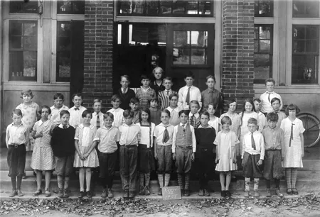 Alice Anderson McMaster with her students at the Clifton School, 1928 — a glossy black-and-white class photograph. Alice stands in the doorway at the back; some three dozen children are ranged on the school steps in front of the brick building.