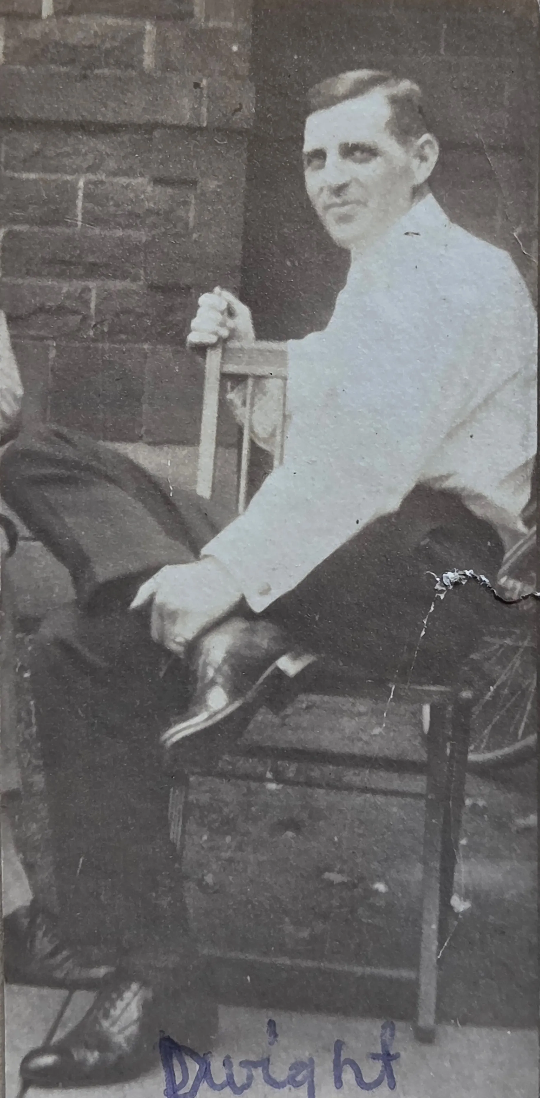 Dwight Kennedy Chenoweth as a young adult, c. 1910-1920, seated cross-legged in a wooden chair against a brick wall (likely a porch). He is in dark trousers, white dress shirt, and a thin tie; smiling slightly at the camera. Blue-ink caption "Dwight" handwritten at the bottom of the print, in the family-album hand. From Roberta Burnes Walker's keeping.