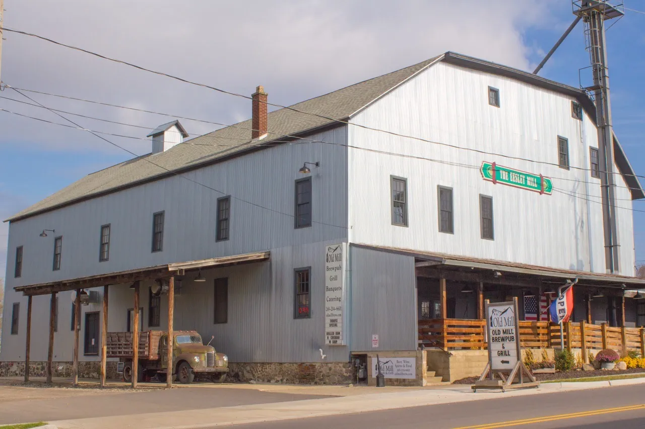 The Eesley Mill at 717 E. Bridge Street, Plainwell, Michigan, present day — the joined mill and elevator under vertical metal siding, gambrel roof at right with the elevator leg above it, gable roof and cupola at left, a brick stack, and a posted loading-platform overhang running the length of the street side. A green sign on the upper wall reads THE EESLEY MILL; Old Mill Brewpub & Grill signage stands at the entrance, with a vintage flatbed truck parked under the overhang.