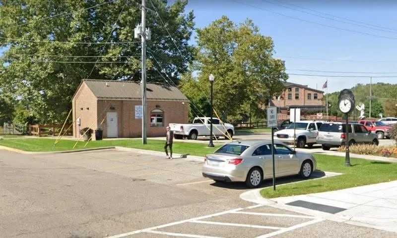 The west side of N. Main Street, Plainwell, in 2019 — a small brick utility building, a parking lot with cars, a street clock, and brick storefronts beyond.