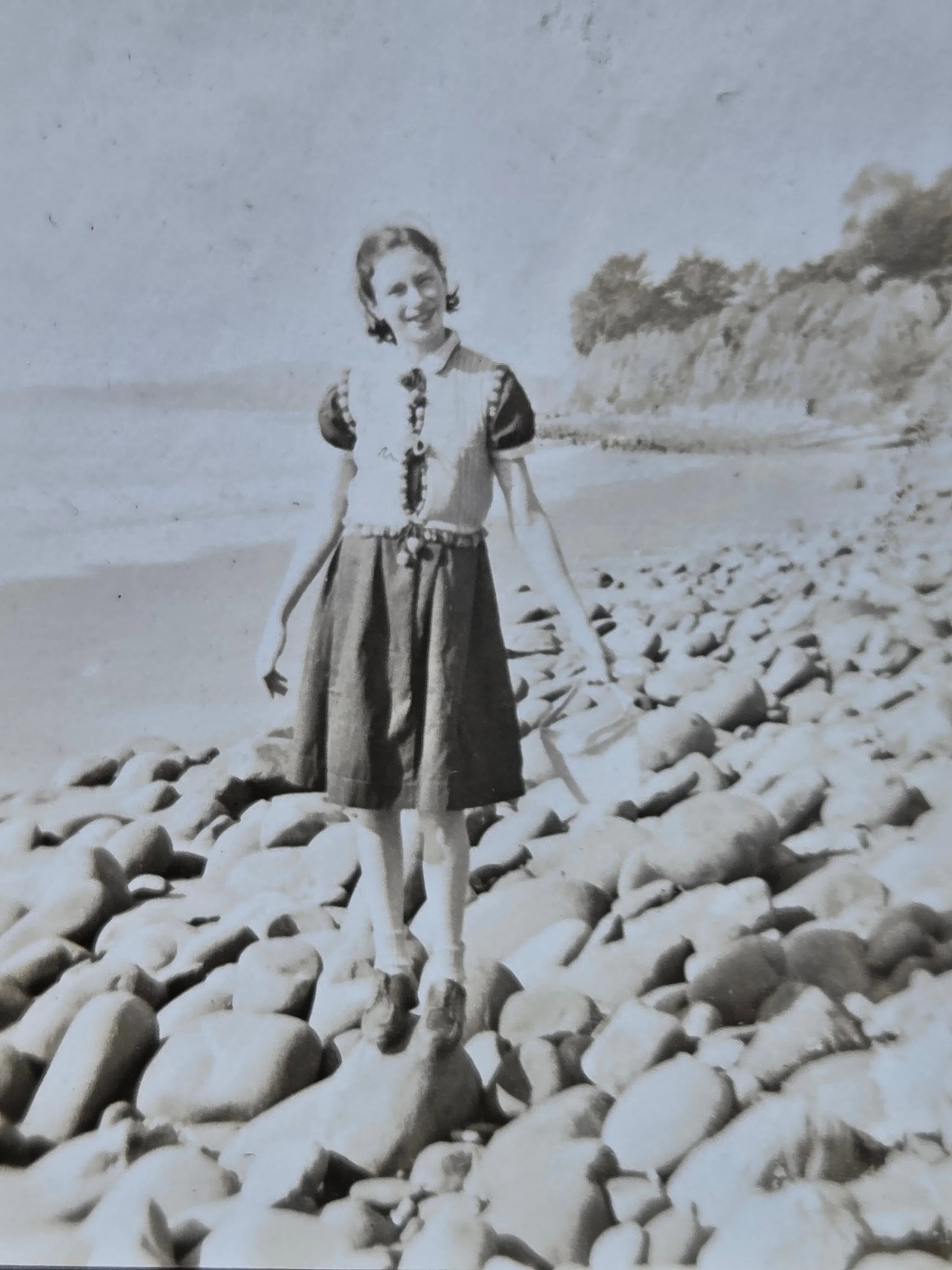 Helen Eesley as a girl of about twelve, standing on a pebble-and-cobble beach with the Pacific Ocean behind her, smiling at the camera. She is in a light cap-sleeved blouse with decorative trim and a long dark skirt, white socks and oxford shoes; her hair is set in shoulder-length waves. To the right of the frame: a wooded shoreline; to the left: open water and a flat horizon. Winter 1936, California. From Roberta Burnes Walker's keeping — taken on Helen's westbound trip with her mother Lillie Dale Chenoweth Eesley.