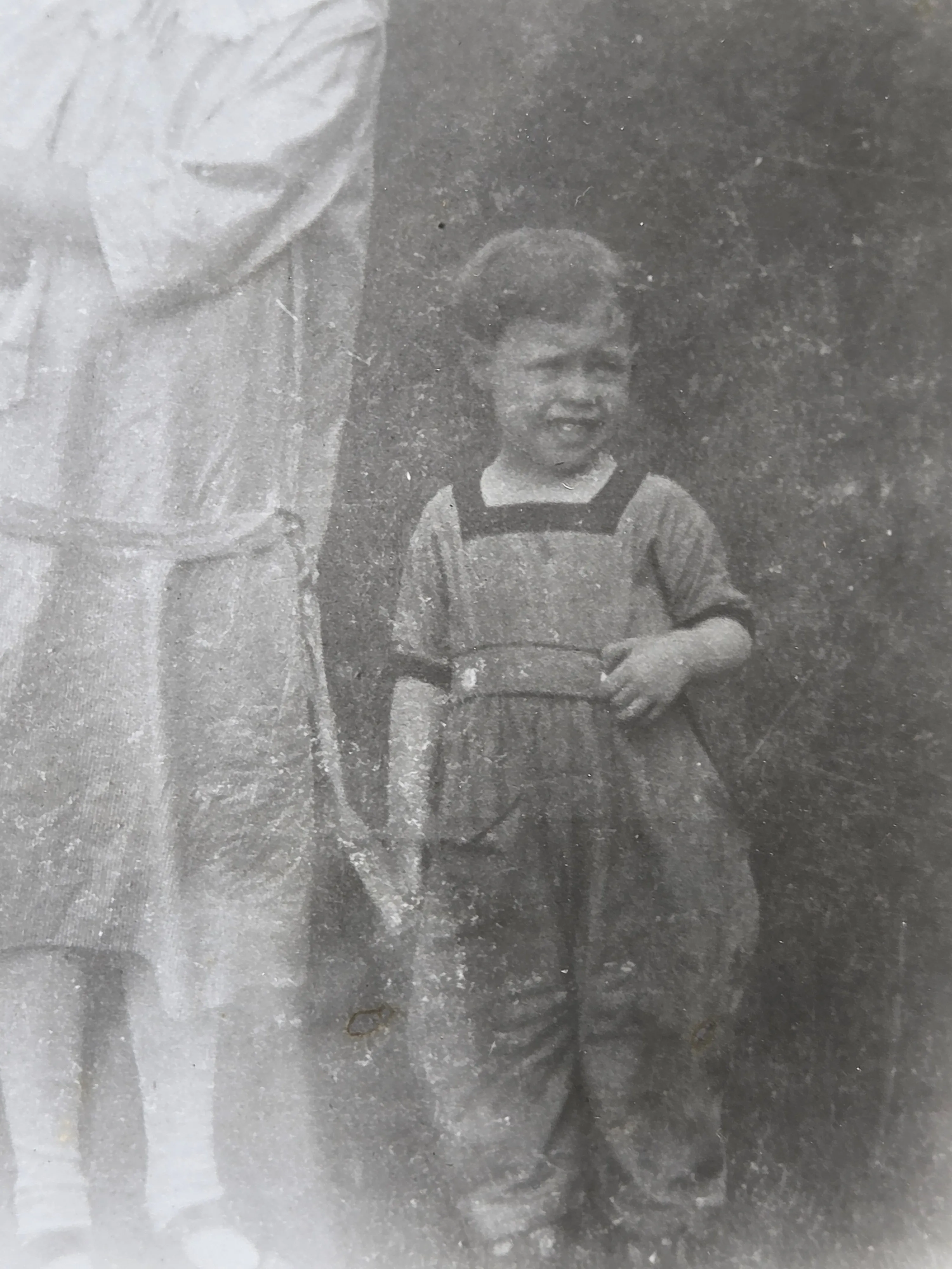 A young child of about two or three years old in dark overalls and a light shirt, smiling at the camera; cropped at left to a partial blurred figure (originally Aunt Mary Eesley Bean holding the photograph's other small child). Roberta Burnes printed this in high school from negatives she found in her mother Helen's family papers. c. 1923-1924, location not recorded — likely Bexley or Grove City, Ohio. The only known solo photograph of Jean Goldie Eesley.