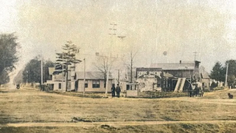 An early hand-tinted view across the open triangular Flat Iron ground at Plainwell, Michigan — bare grass in the foreground, frame houses, a small shed, utility poles and two figures standing at the street line behind.
