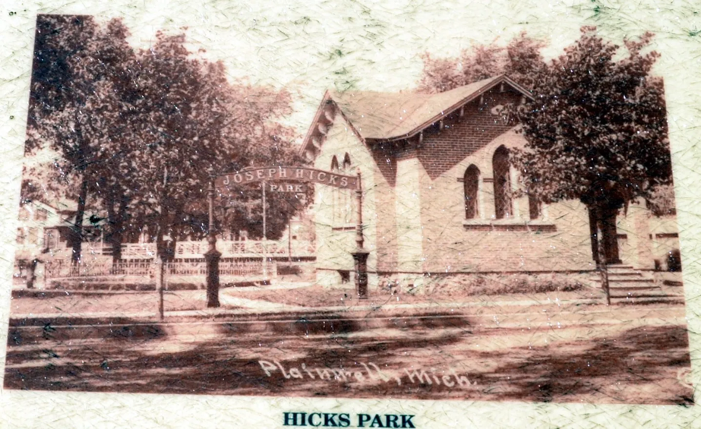 A sepia postcard reproduction captioned “Plainwell, Mich.,” showing an iron entrance arch lettered JOSEPH HICKS PARK over a walk, with a brick gable-fronted church and a low fence beside it; printed caption HICKS PARK below.