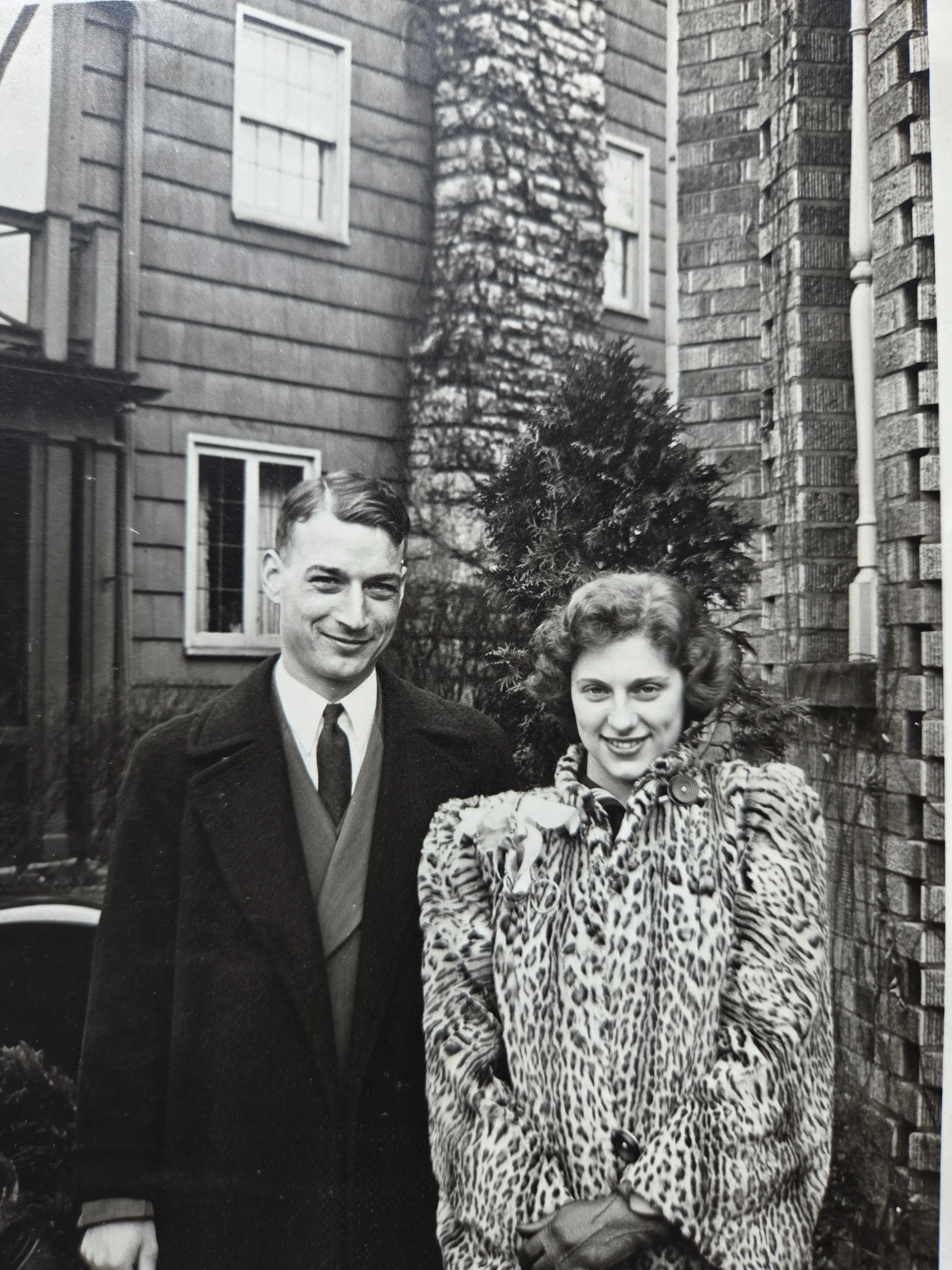 Lyle Eesley with the young woman he would have married had he not died at Cabanatuan in 1942 — Lyle in dark coat and tie, smiling at the camera, his arm at her shoulder; she in a leopard-print fur coat with a brooch at the collar, fashionable c. late 1930s. Outdoors in front of what may be the Bexley house. c. late 1930s. From Roberta Burnes's keeping via Helen Eesley Burnes; identification of the young woman pending — Chuck and Roberta to confirm.