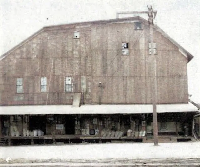 The Sunshine Flour Mill from its loading-dock side, early 1900s — the long covered platform along the ground floor stacked with sacks of flour, the mill rising behind.