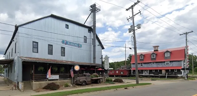 A 2019 Google Street View of the Eesley Mill from across East Bridge Street — the grey mill with its covered porch at left, and to the right the neighboring red-roofed barn and a red railroad caboose beside the tracks.