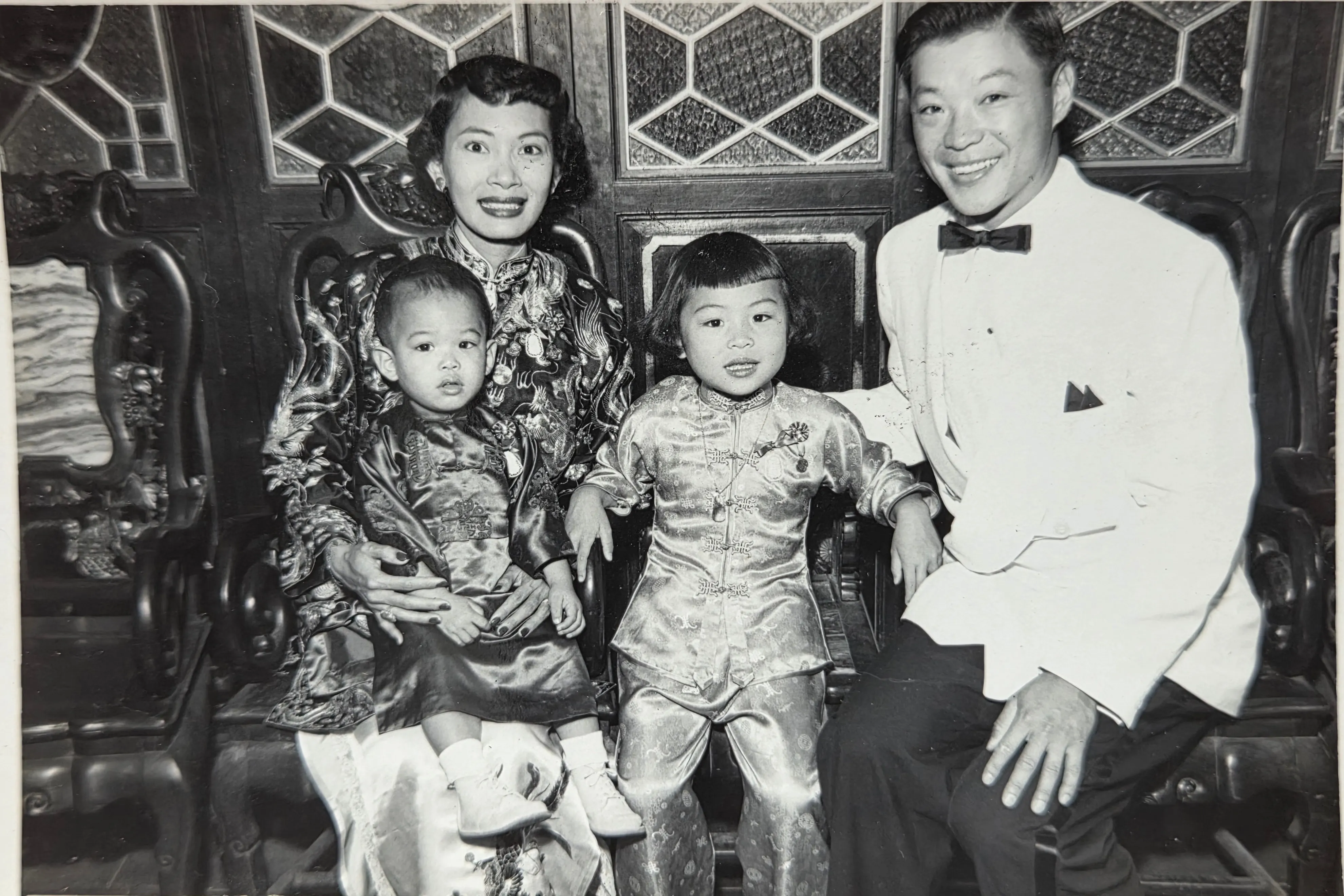 Stella, Ted, SueLynn (right), and Glenn (?, left) in a mid-1950s formal portrait at what reads as a Chinese-restaurant or community-association hall. Stella in a brocade cheongsam, Ted in white dinner jacket.